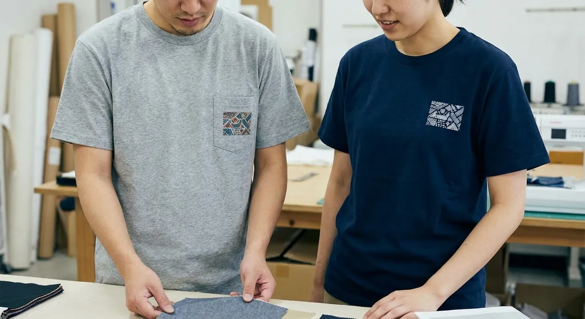 Grey and navy t-shirts with a small square print on the chest, being compared on a worktable