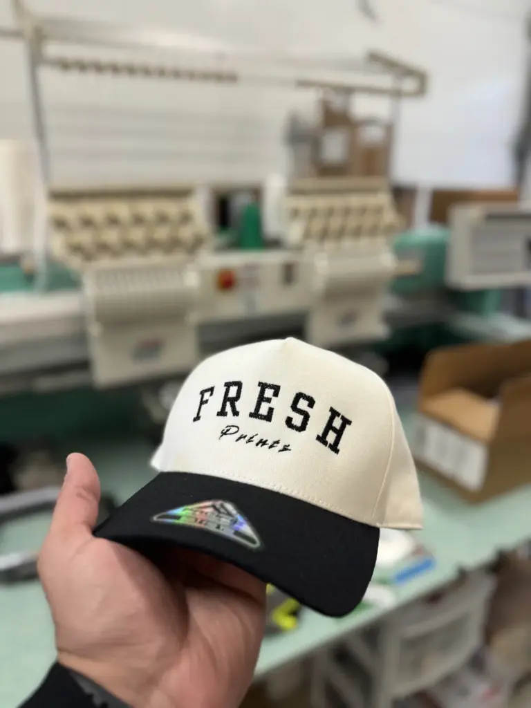 Cream and black Fresh Prints embroidered cap held in front of the embroidery machines in our Surrey shop