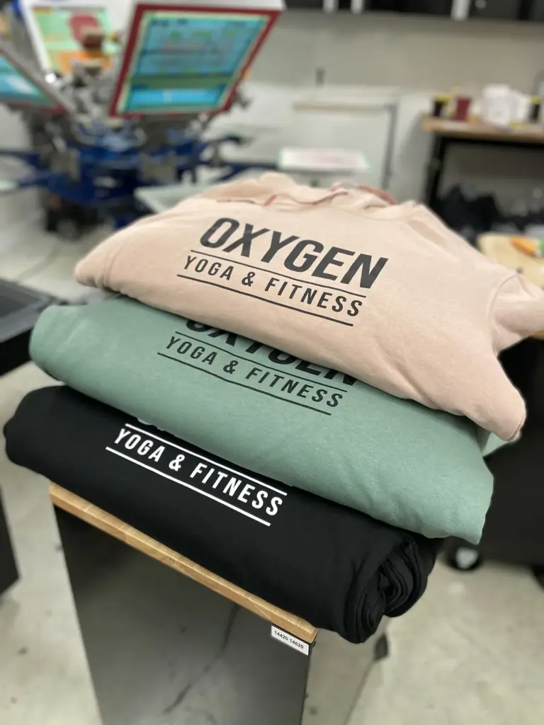 Stack of pink, green and black hoodies printed with the Oxygen Yoga & Fitness logo