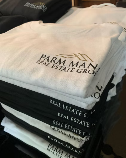 Folded white and black t-shirts printed with the Parm Man Real Estate Group logo